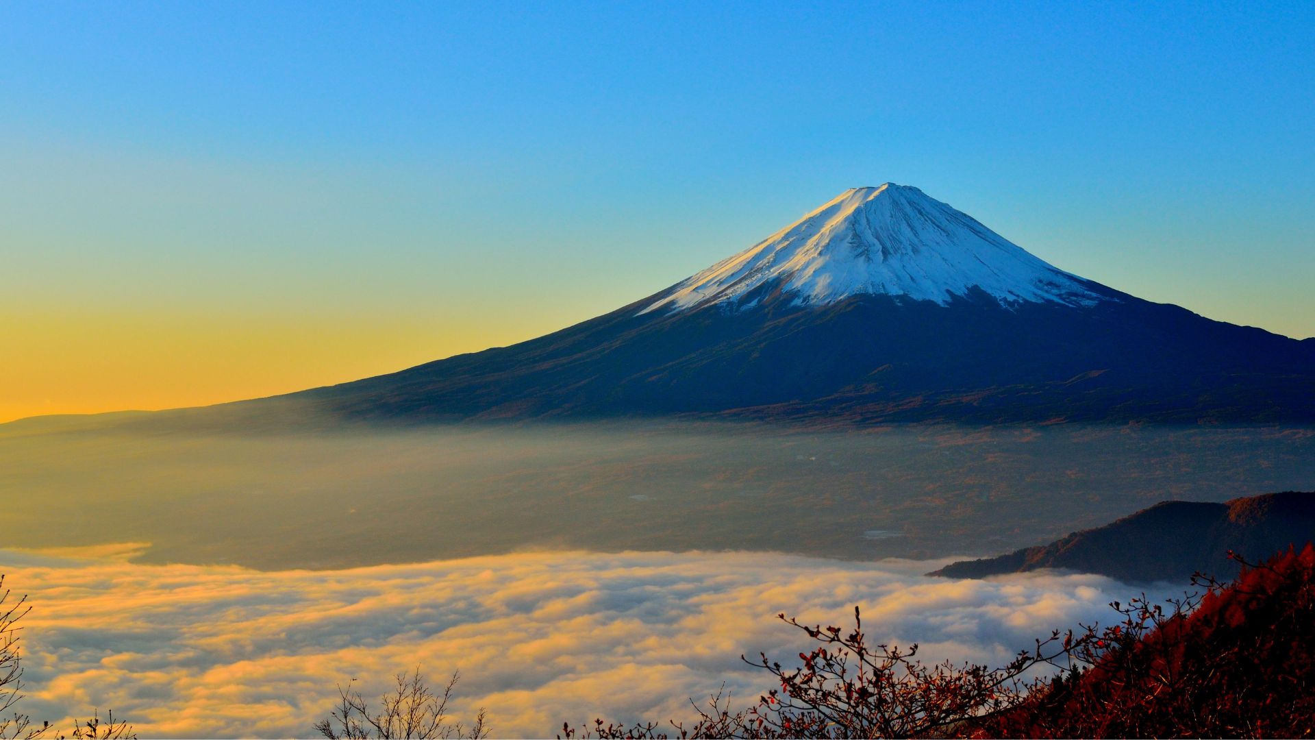 Mont Fuji imposant avec une petite montagne au premier plan. Ciel bleu vif avec des nuages jaunes et blancs en bas. Arbres de couleur rouge en haut de l'image avec le mont Fuji en arrière-plan.